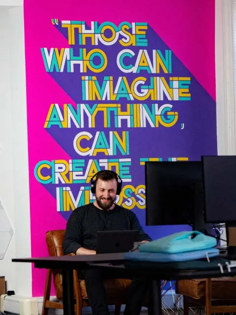 One of our producers sitting at his laptop infront of a mural that reads 'Thos who can imagine anything, can change the world'